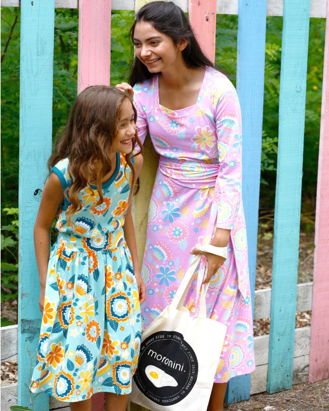 Mädchen in moromini Outfit aus Biobaumwolle vor buntem Zaun